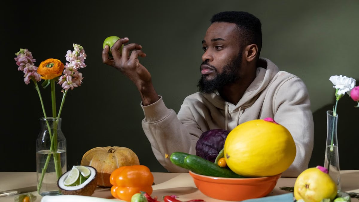 A man in a hoodie surrounded by fresh produce and flowers, contemplating a green apple.